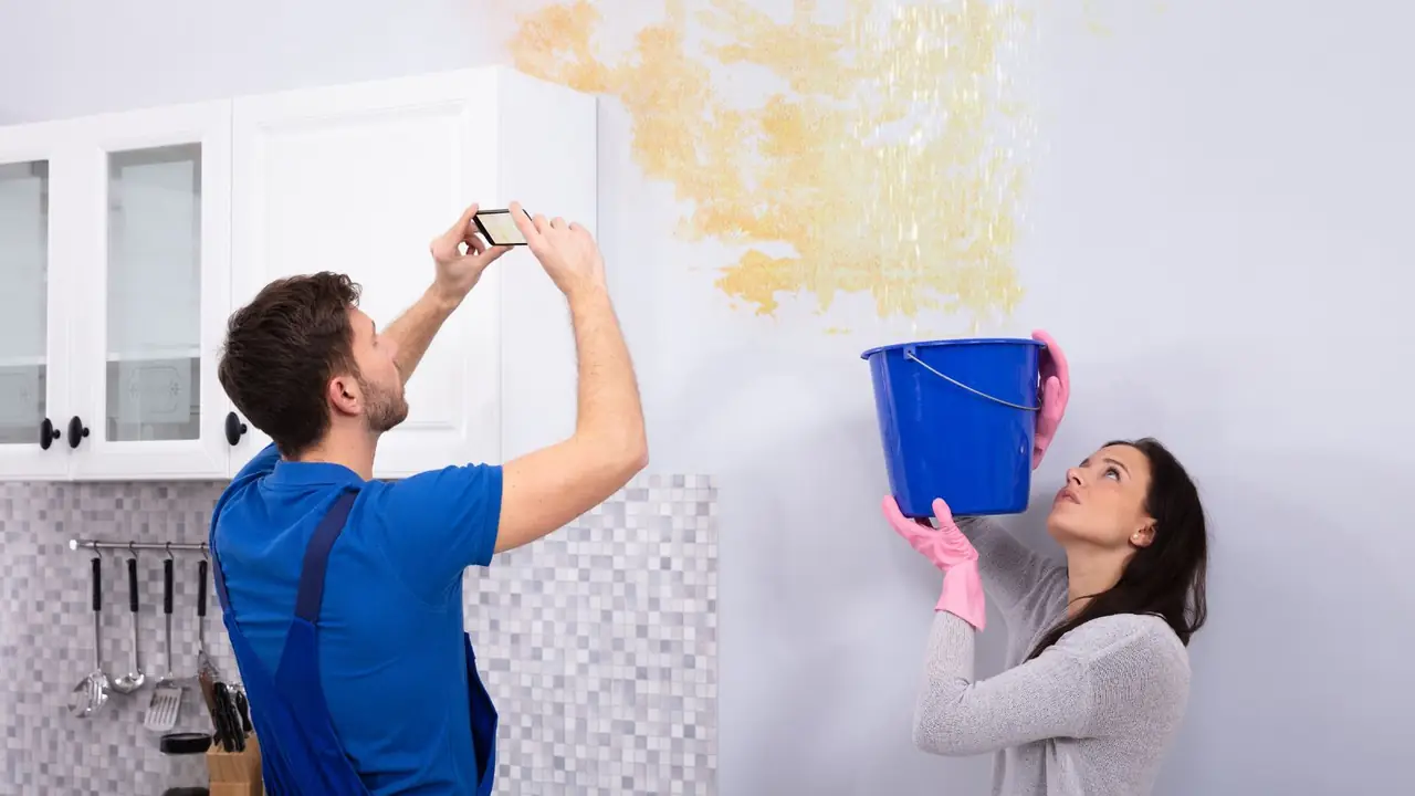 A man photographing a water-stained wall while a woman catches a leak in a bucket